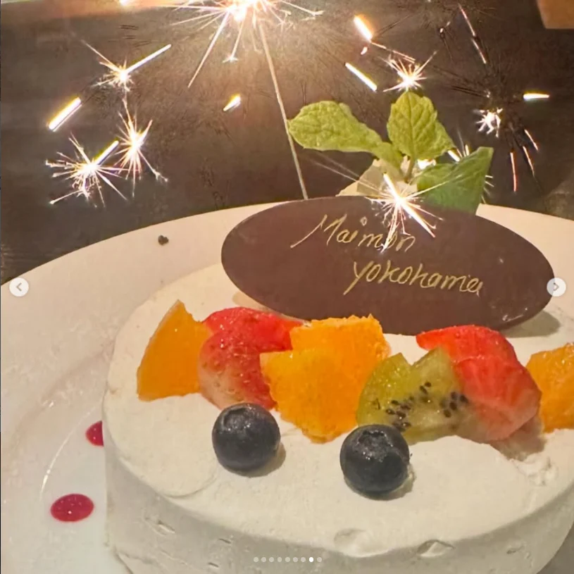 A fruit-topped white cake with a lit sparkler and a chocolate plaque.