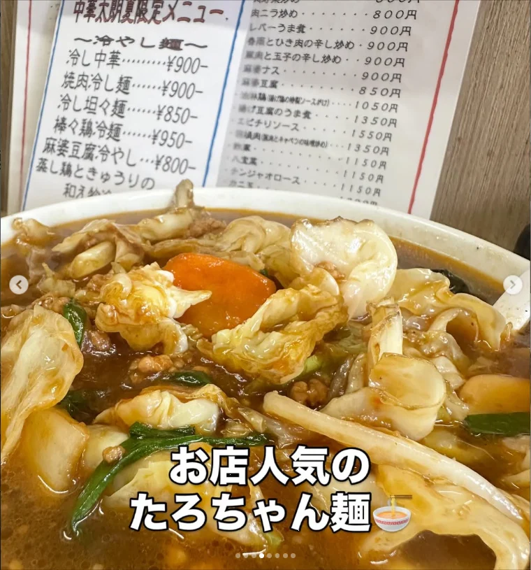 A bowl of noodles with golden fried pieces, greens and an egg, with a menu behind it.