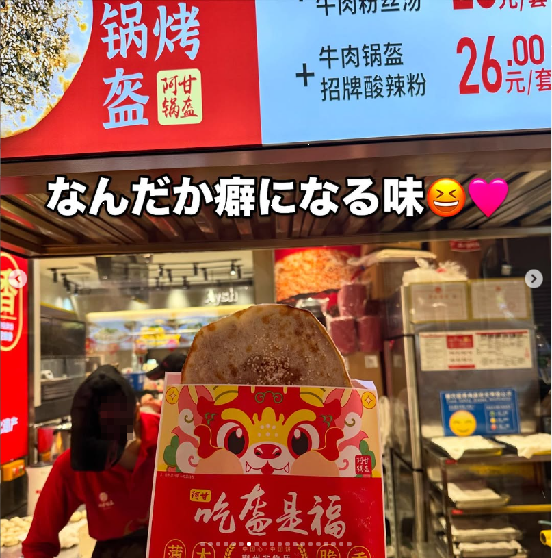 A round baked snack in a paper sleeve at a food stall beneath menu boards.