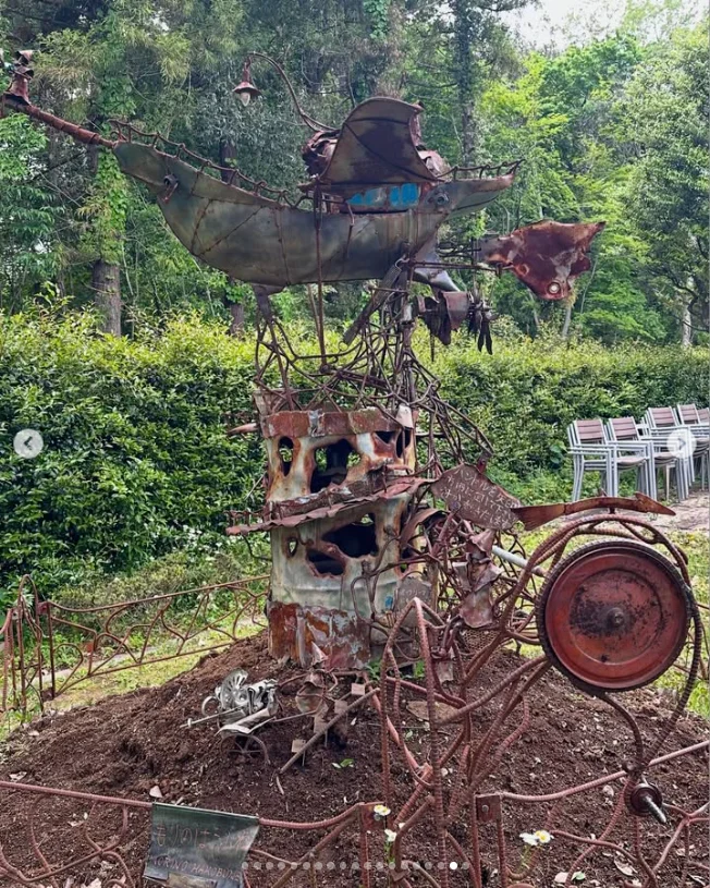 An outdoor metal sculpture of stacked objects and wheel-like forms among trees.