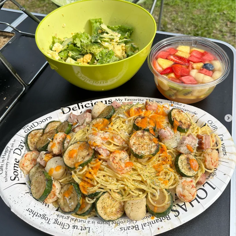An outdoor meal of pasta and browned vegetables beside bowls of cut vegetables.