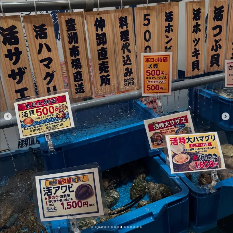 Blue tanks beneath wooden Japanese menu boards showing shellfish names and prices.