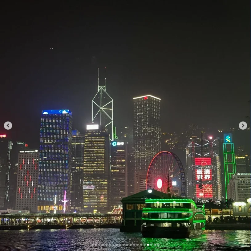 A green boat in front of an illuminated waterfront skyline at night.