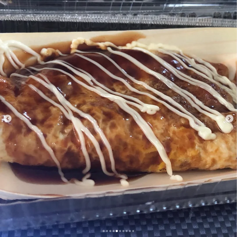 An oblong browned dish drizzled with dark and white sauces in a takeaway tray.
