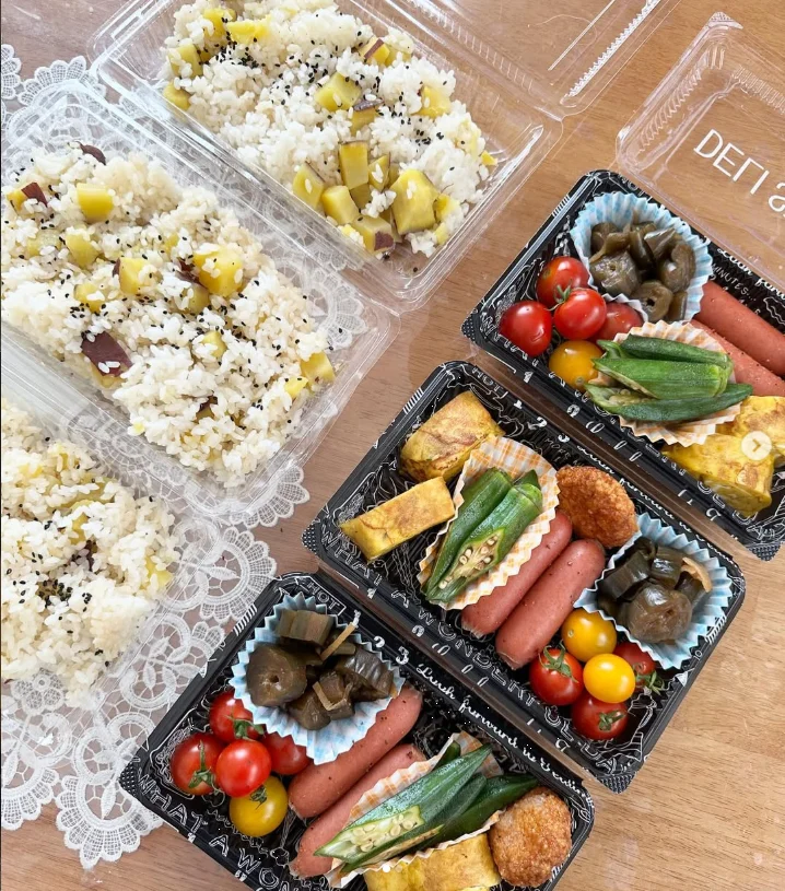 Open lunch containers with rice, sausages, rolled egg, green vegetables and cherry tomatoes.
