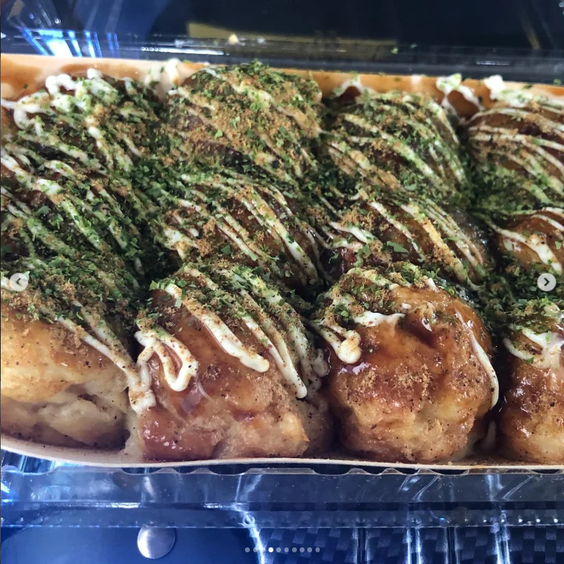 Round browned snacks covered with green flakes and drizzles of pale and dark sauce in a takeaway tray.